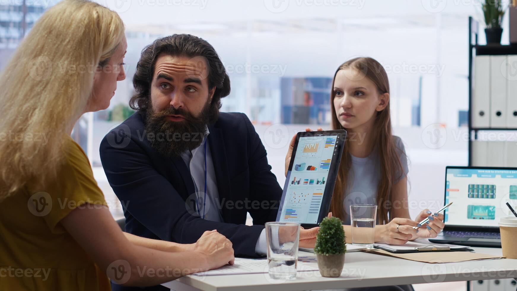 Financial advisor showing statistical graphs on tablet to clients, analyzing market trends to optimize their income. Mother and daughter checking investment plan on device, camera B photo