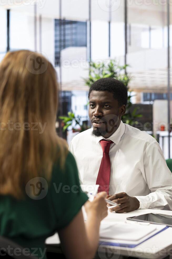 Headhunter tasked with hiring best talent to fill open company position, interviewing man. Employer hired to assess skills and qualifications of african american candidate, doing meeting in office photo
