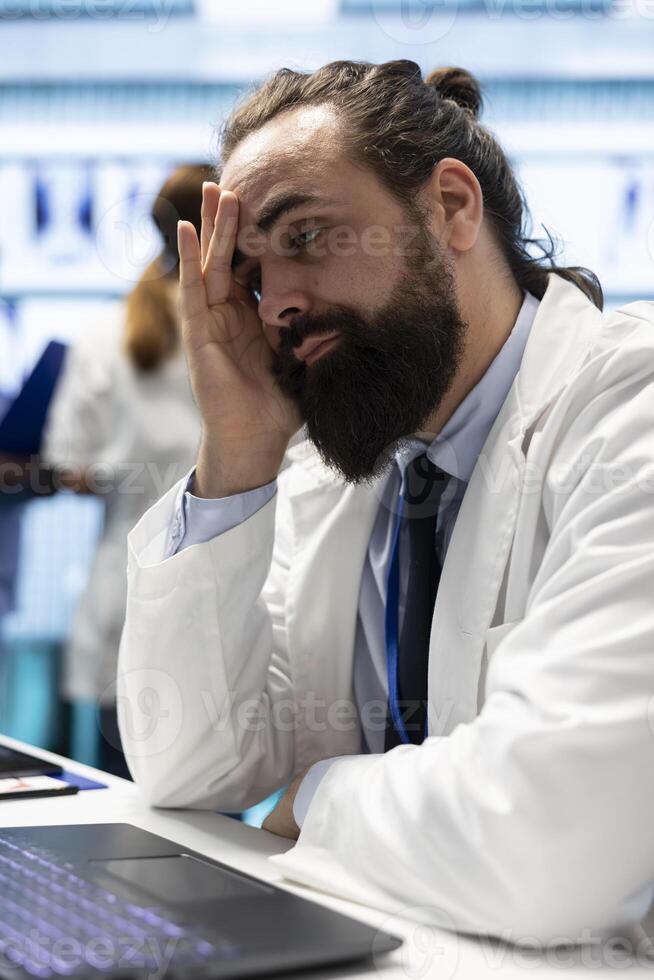 General practitioner feeling tired and stressed after analyzing medical records and test results, ensuring recovery in treatment center. Physician working under pressure to find a cure for illness. photo