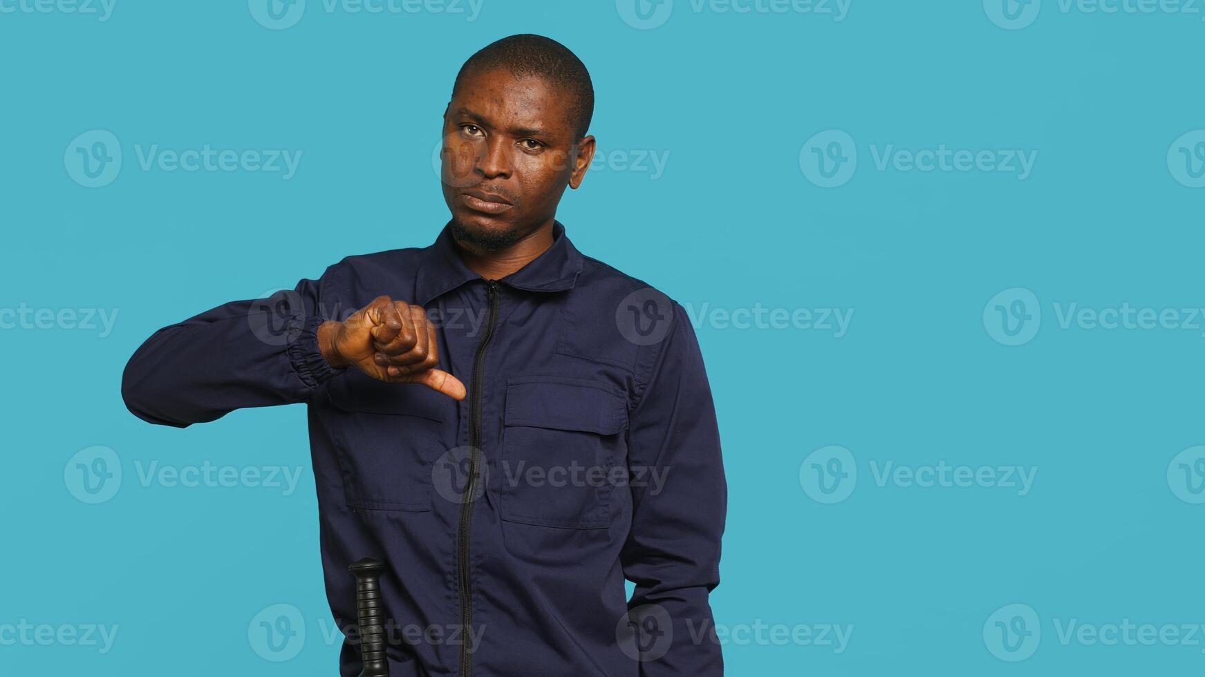 Portrait of security guard doing thumbs down sign gesturing, denying access while overseeing premises. Private law enforcement worker doing negative hand gesture, studio backdrop, camera A photo