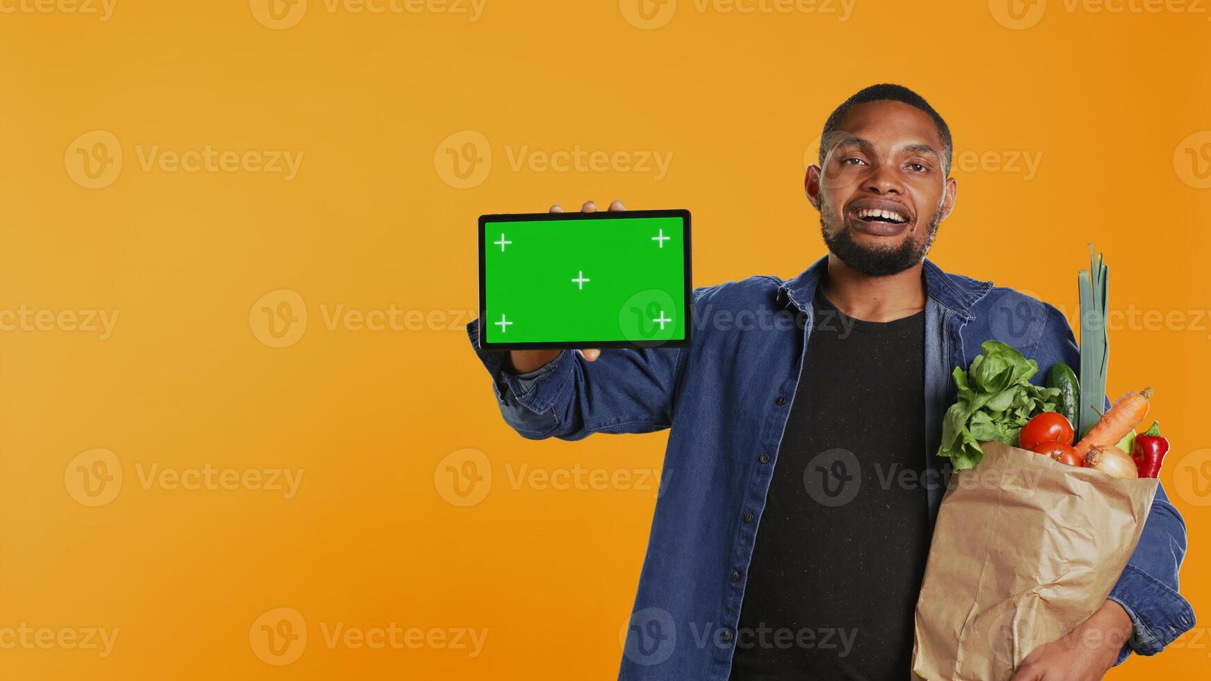 African american guy showing a tablet with greenscreen layout, going on a grocery shopping spree and supporting local farming. Young man advertises an isolated screen and healthy nutrition. Camera B. photo