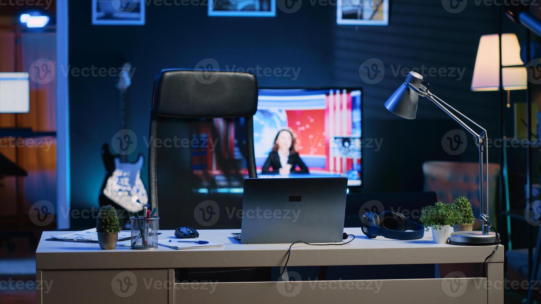 Focus on office desk in front of smart TV in blurry background displaying news program broadcasting. Laptop on table and television screen in empty room showing VOD channel, camera A photo