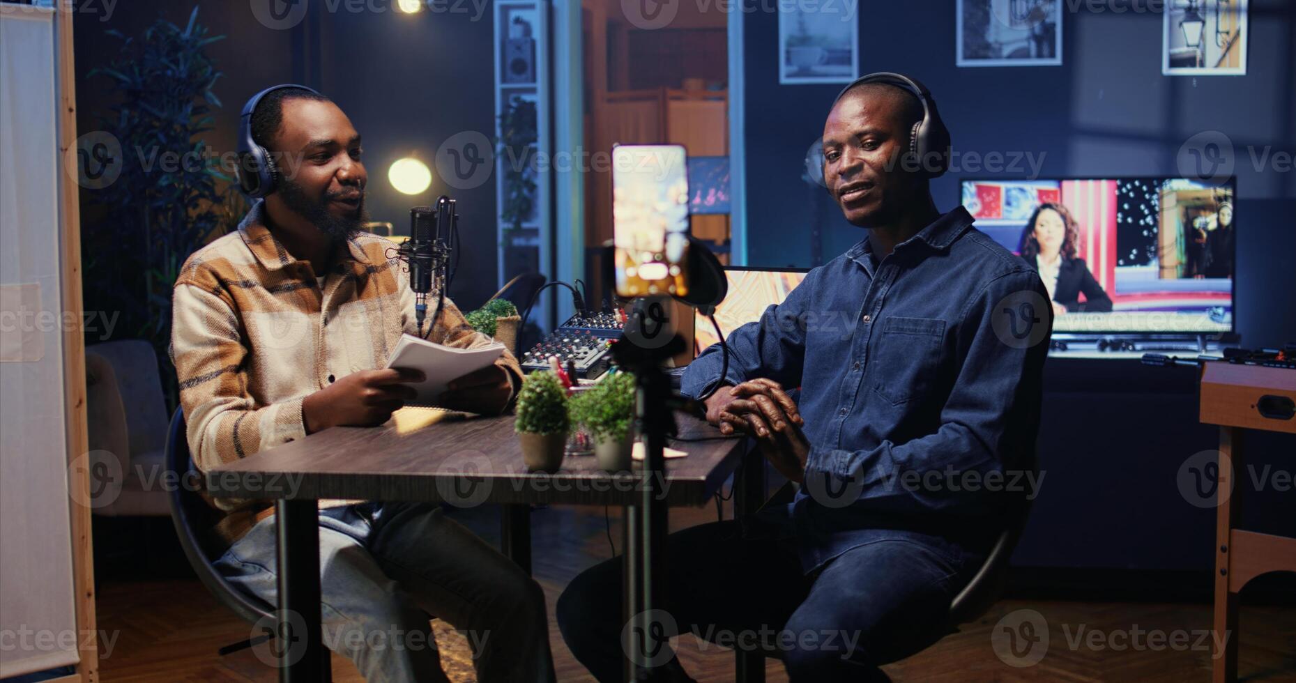 Show host talking with guest in studio, listening to opinion on current events, filming for social media using smartphone on tripod. Influencer recording podcast with mobile phone photo