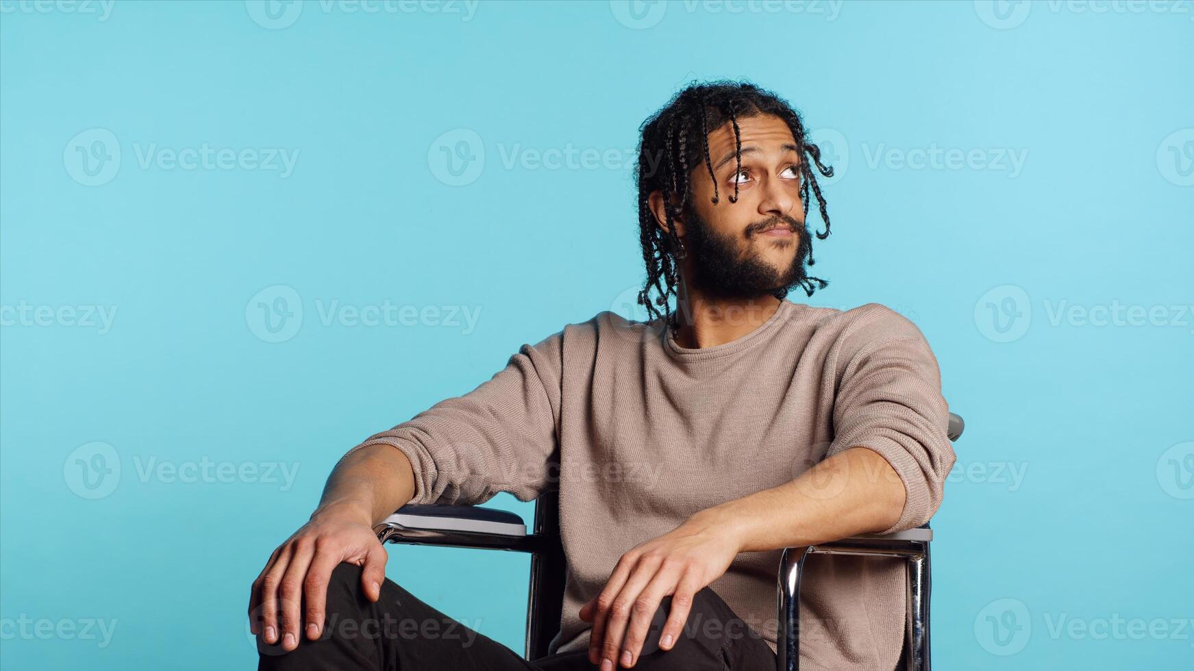 Bored Middle Eastern man with disability in wheelchair impatiently tapping fingers on knee, waiting. Annoyed person with paraplegia looking around, isolated over studio background, camera A photo