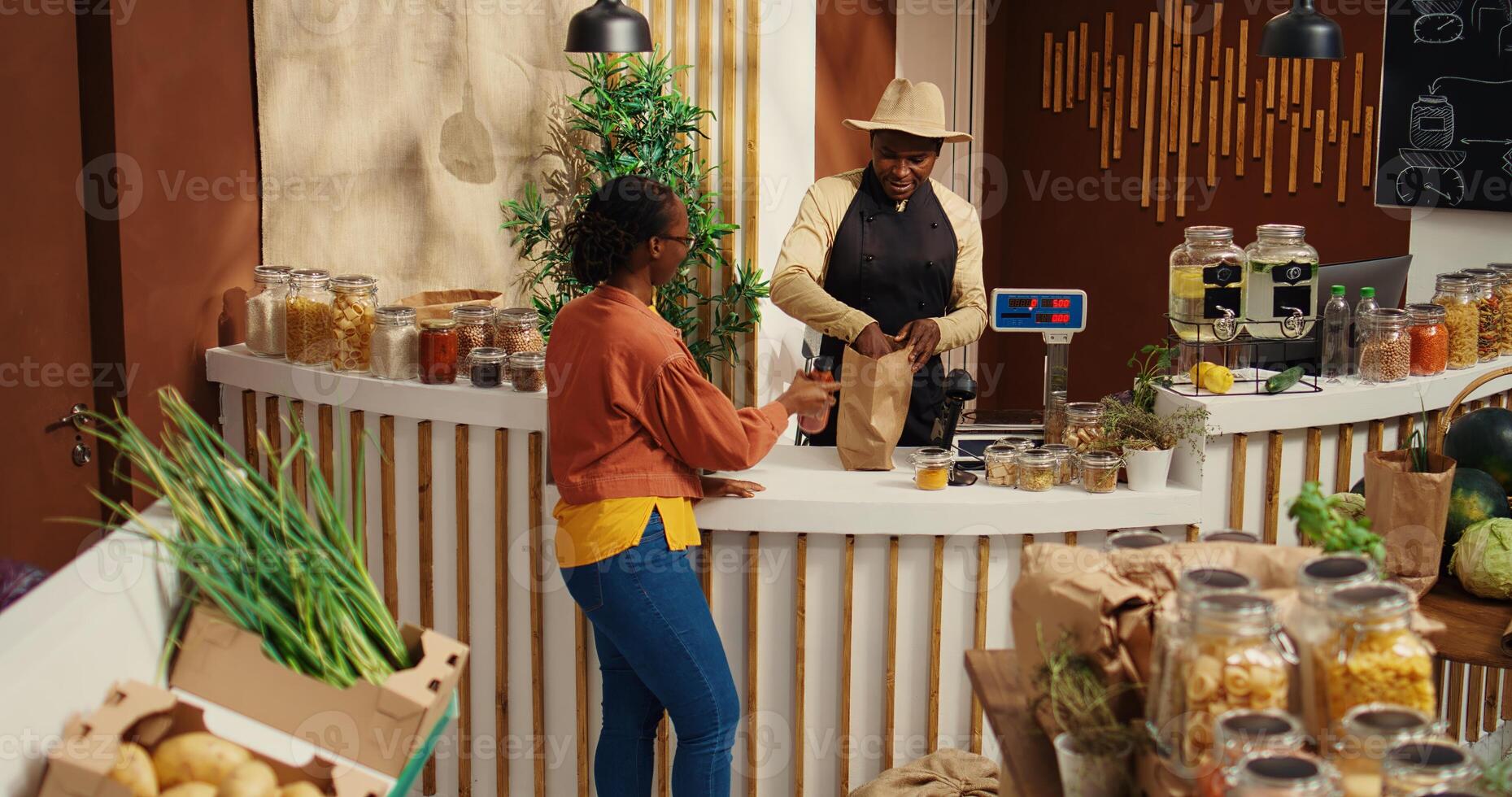 Merchant greeting woman at zero waste eco store checkout, weighting organic produce and bulk items in jars at cash register. African american client buying locally grown natural food. Camera 2. photo