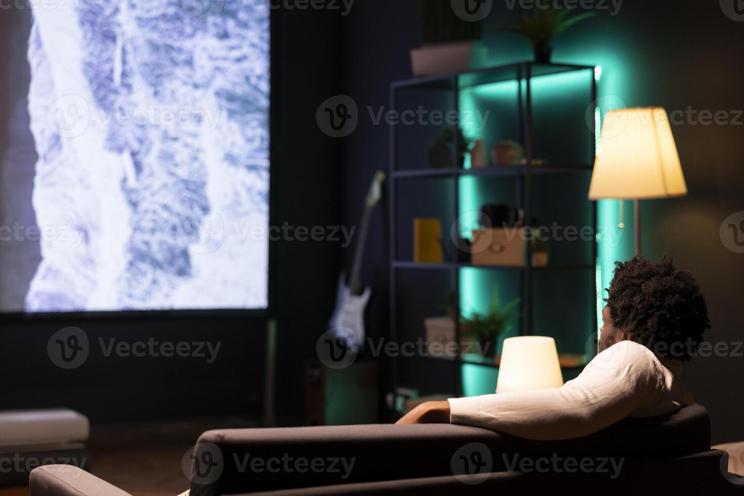 African american man laid up on couch watching nature documentary with breathtaking cinematography on huge TV screen. Person enjoying scenery shots on large television set photo