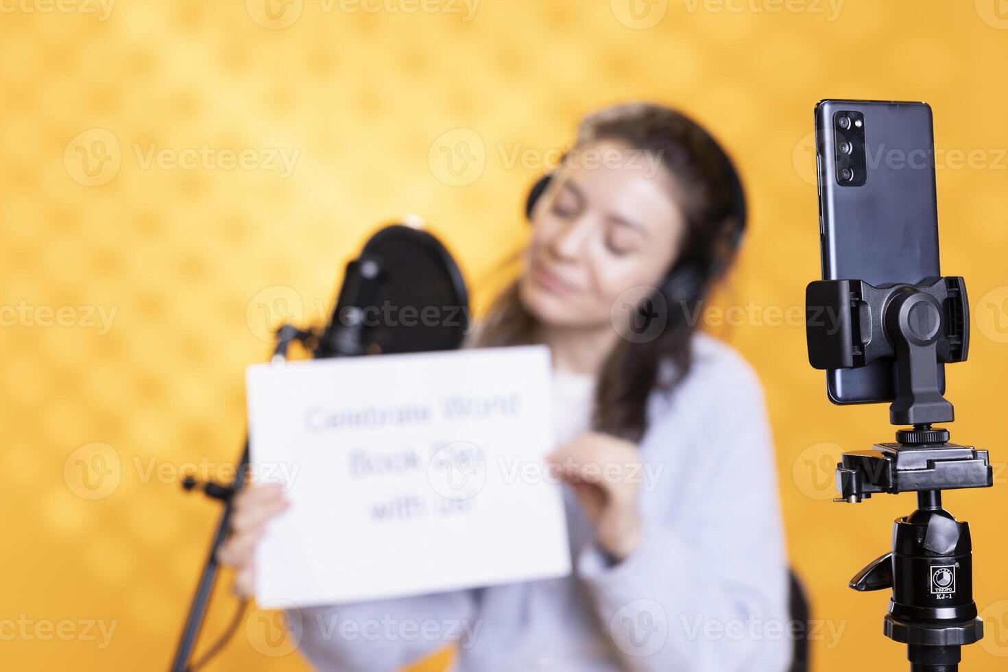 Focus on phone used by woman in blurry background filming promotional for world book day, studio background. Close up on smartphone used by content creator promoting reading photo