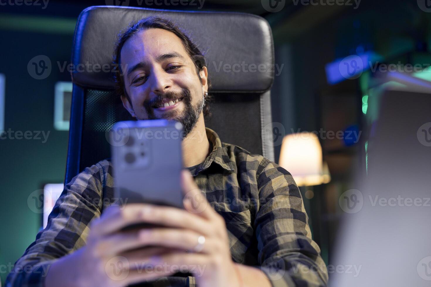 Bearded man sitting in front of laptop holding his cell phone, smiling and browsing the internet. Cheerful young male individual using his mobile device to check his social medias. photo