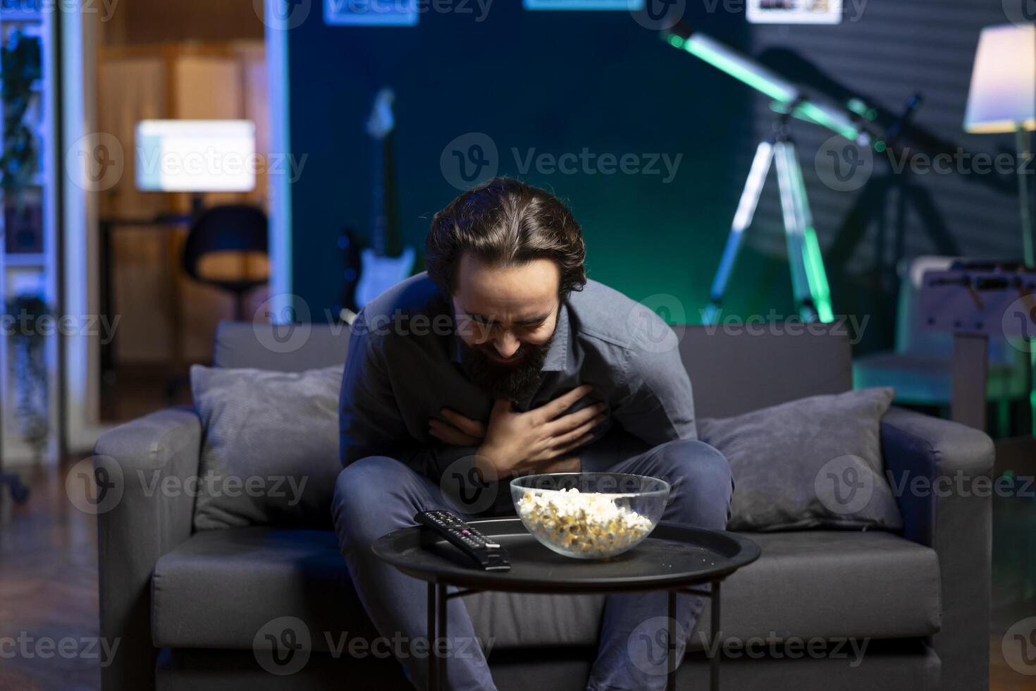 Joyous man sitting on couch having fit of laughter watching funny comedy movie while eating popcorn. Upbeat person in apartment out of breath after laughing too much at hilarious joke in film scene photo
