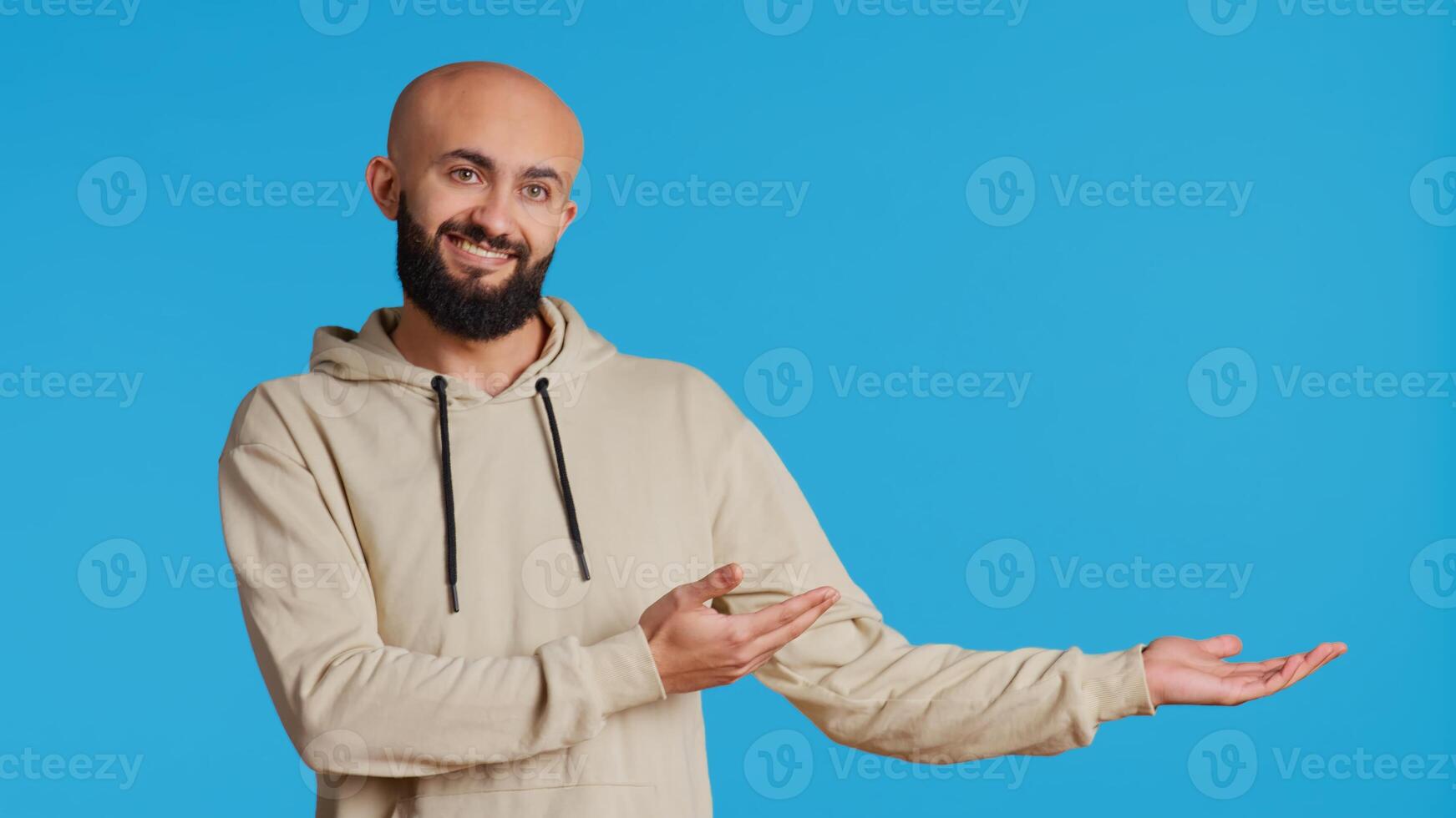 Cheerful man creating ad in front of studio camera, pointing to left or right sides over blue background. Middle eastern person showing commercial or advertisement for marketing campaign. Camera 2. photo