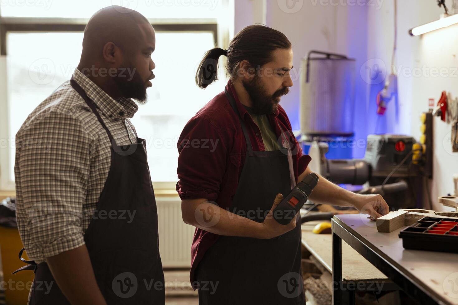 Cabinetmaker teaching apprentice to drill hole into timber block in studio. Woodworking pupil learning from master in joinery how to insert nail into piece of wood using power drill photo