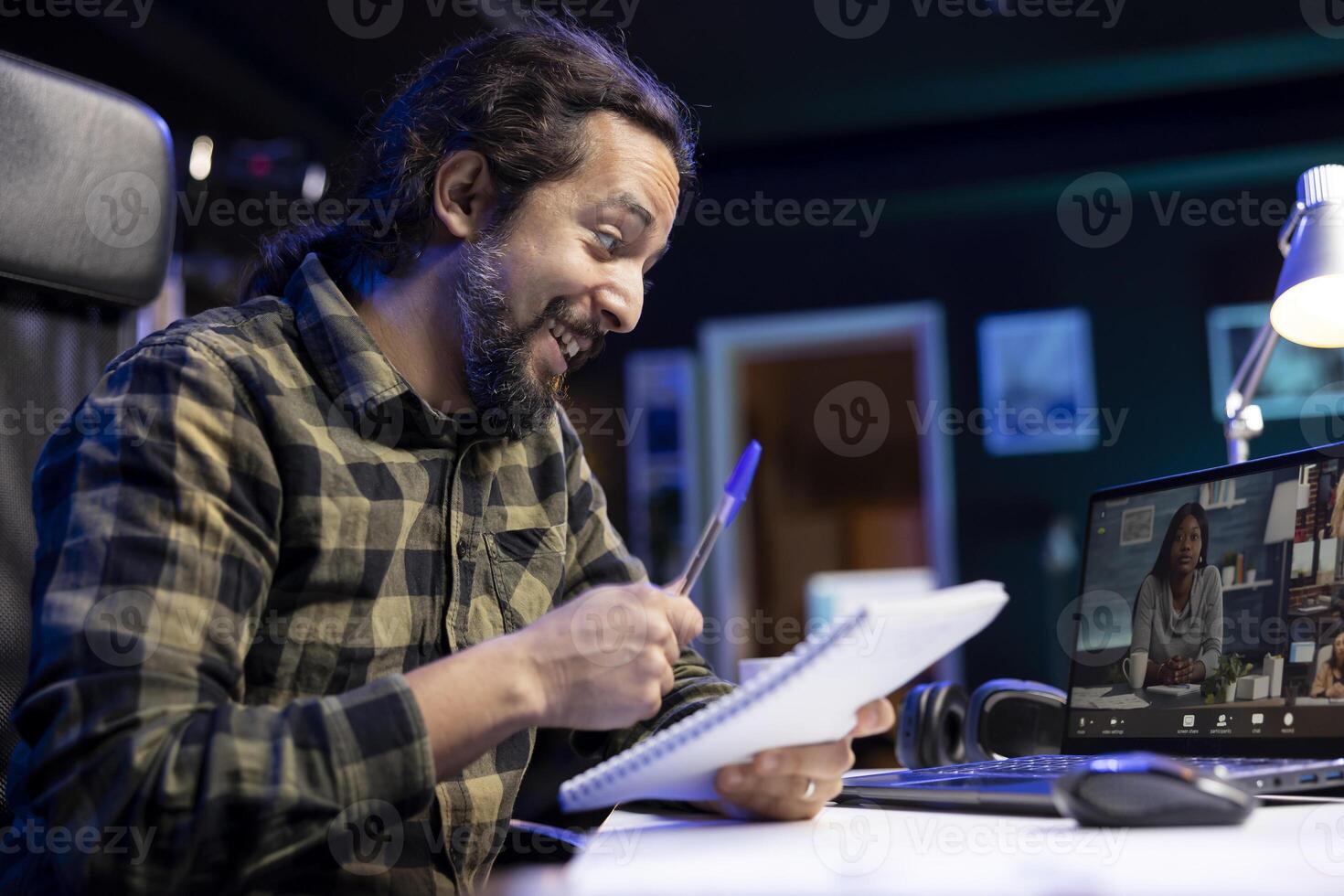 Male college student attending a virtual meeting through his laptop, cheerfully writing down notes. Smiling freelancer with a notebook positively attending conference call. photo