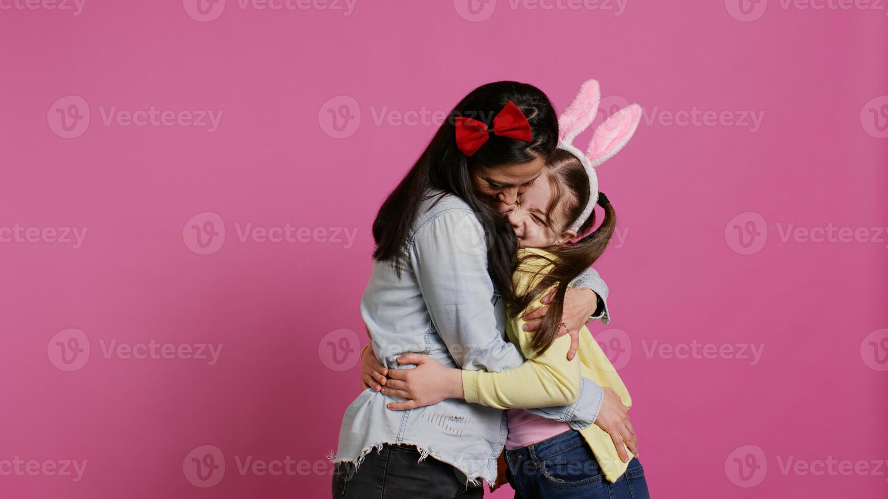 Lovely schoolgirl with bunny ears an her mom waving on camera, having fun and laughing against pink background. Cheerful mother and her daughter embracing and kissing each other. Camera B. photo