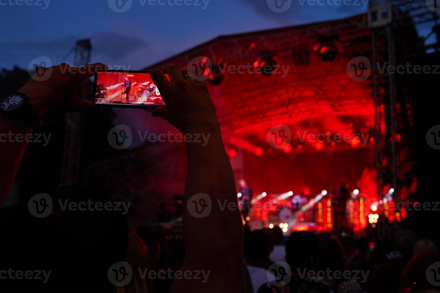 Person standing in front of stage, recording live music concert on smartphone. People crowd near stage enjoys musician band's performance photo