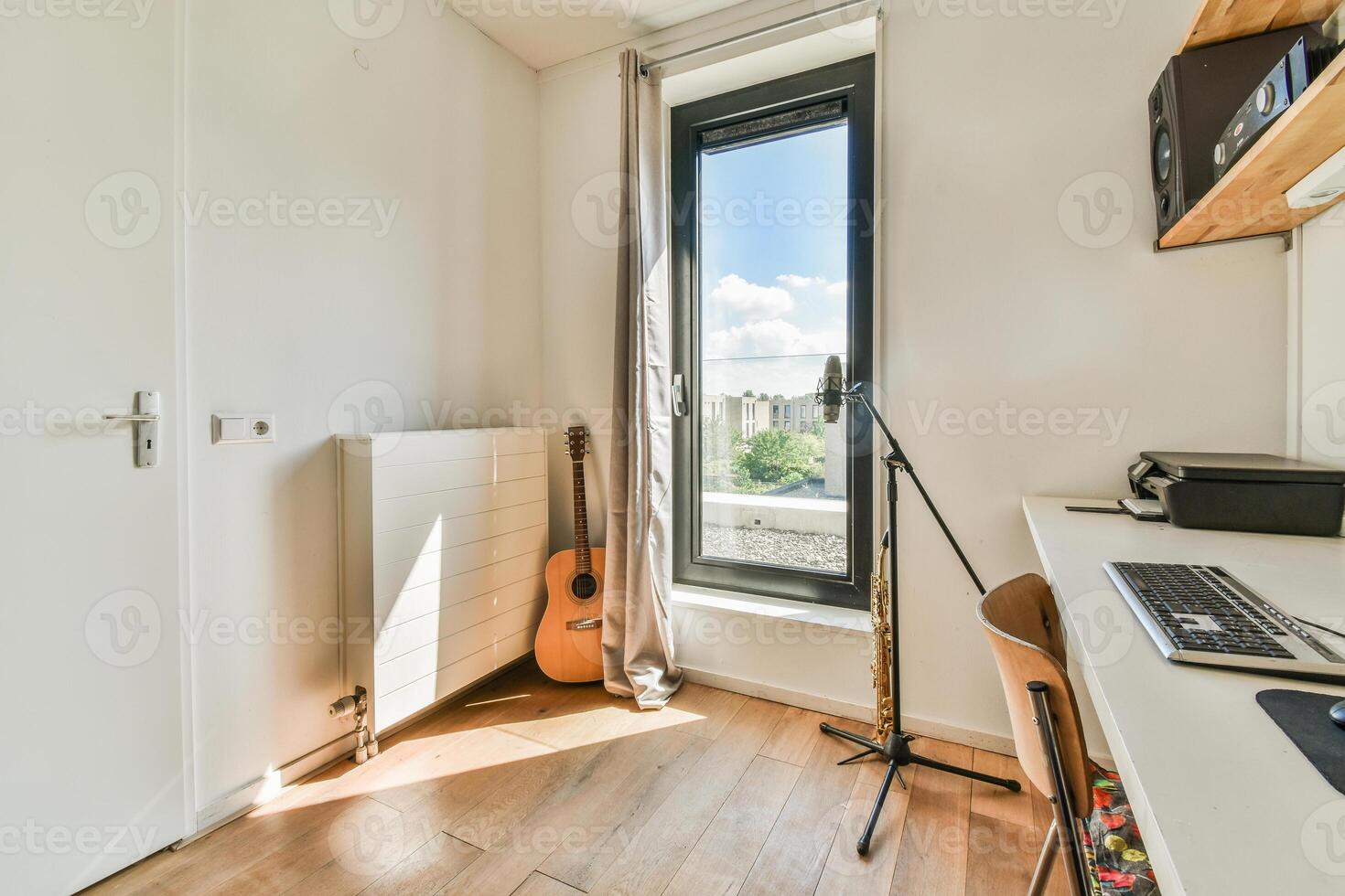 A small room with a guitar, computer and a window photo