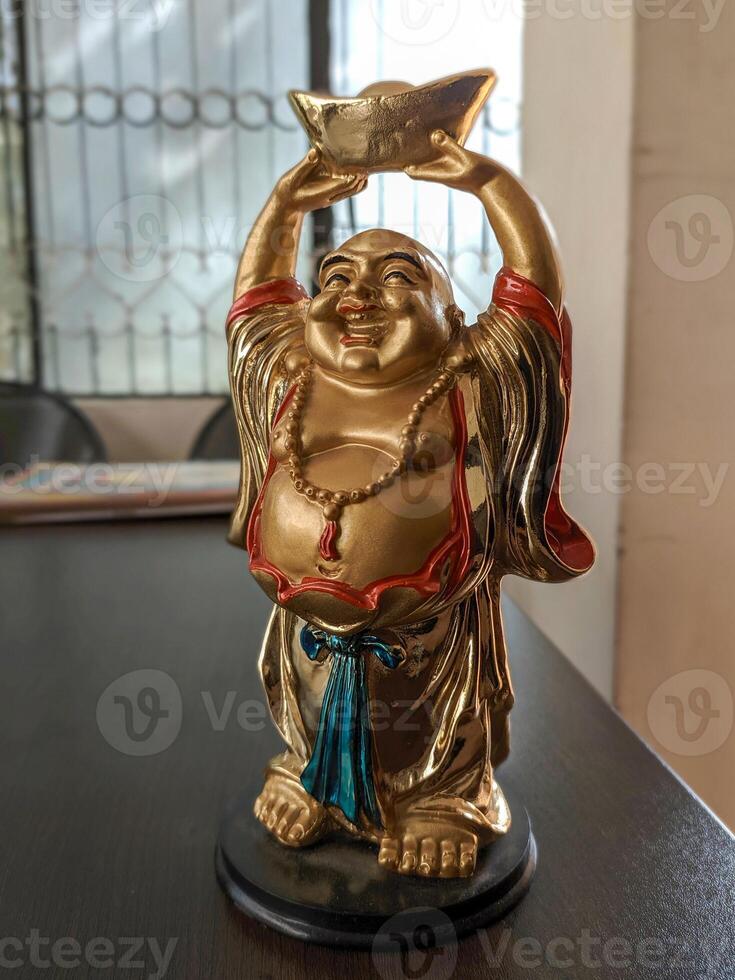 A golden Laughing Buddha in a red robe and blue sash holds a gold ingot, standing on a black base. It is on a dark wooden surface with a blurred, iron grilled window in the background. photo