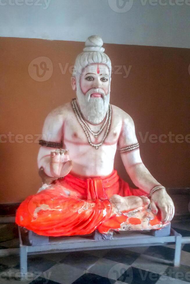 A worn statue of a seated Hindu sage with a white beard, orange dhoti, and beaded jewelry, holding one hand in a blessing gesture. photo
