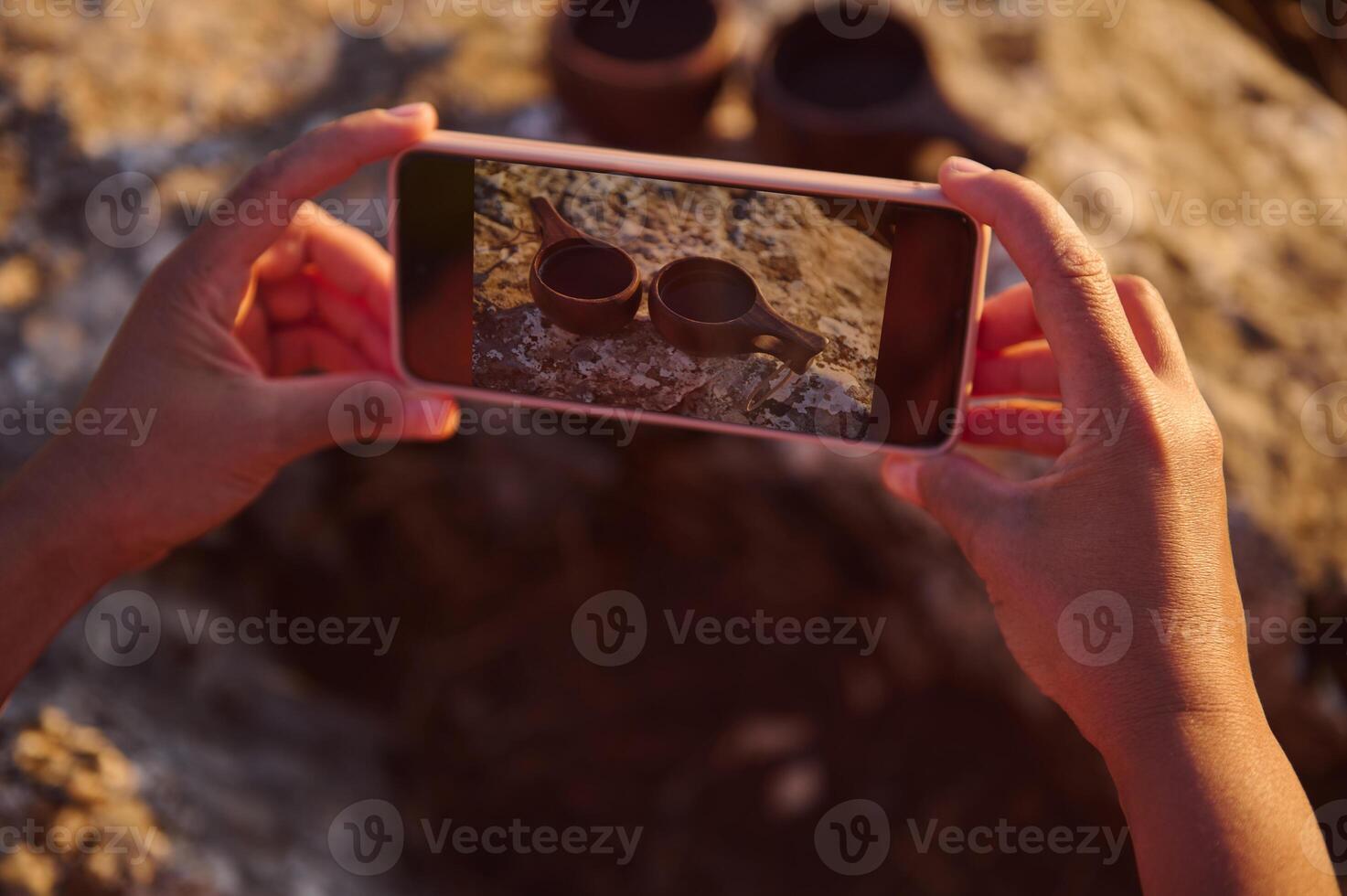 Close-up of hands photographing wooden cups on rocky surface with smartphone in natural light. photo