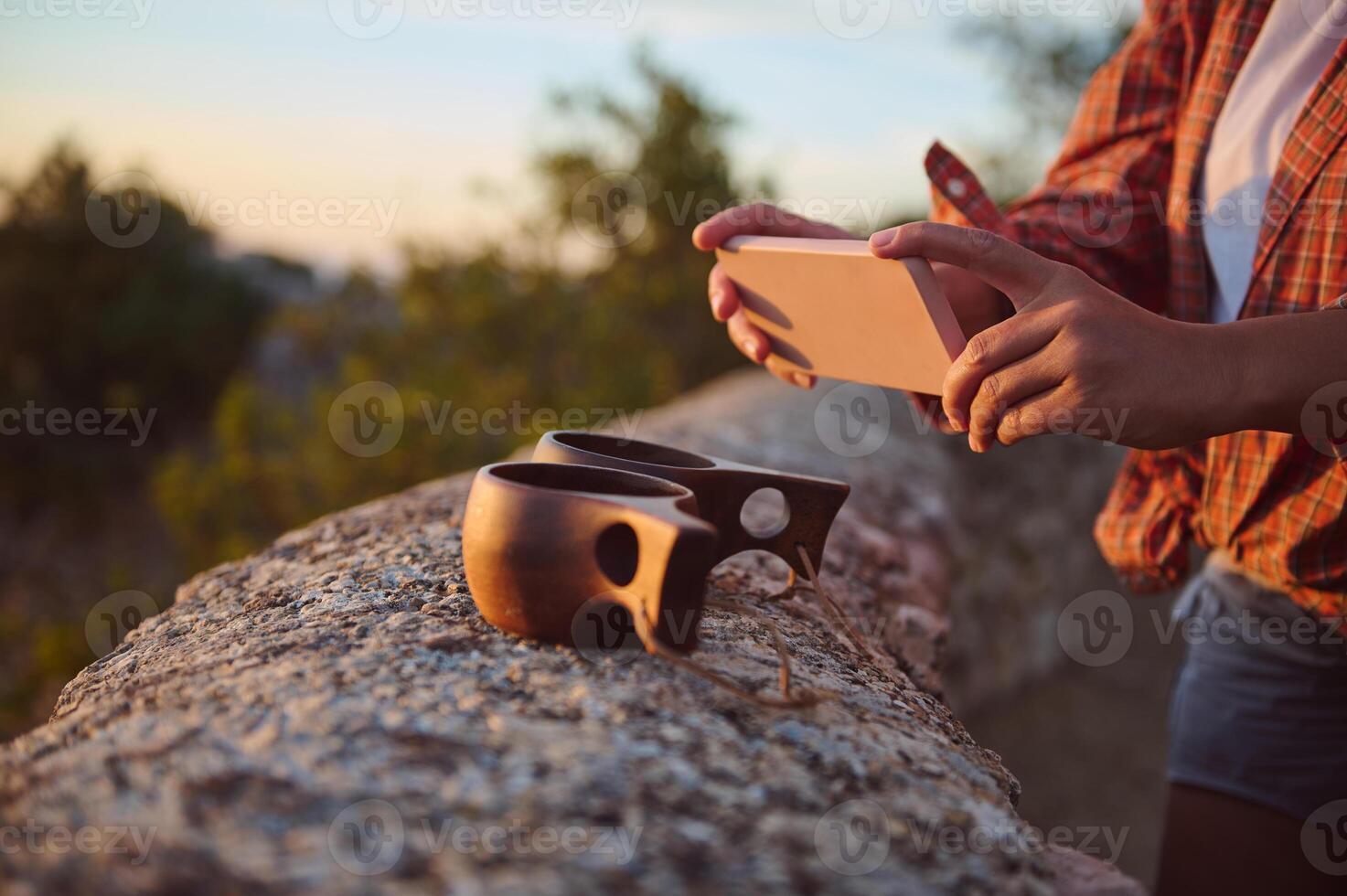 Person photographing wooden mugs with a smartphone on a stone wall during a sunset in the countryside. photo