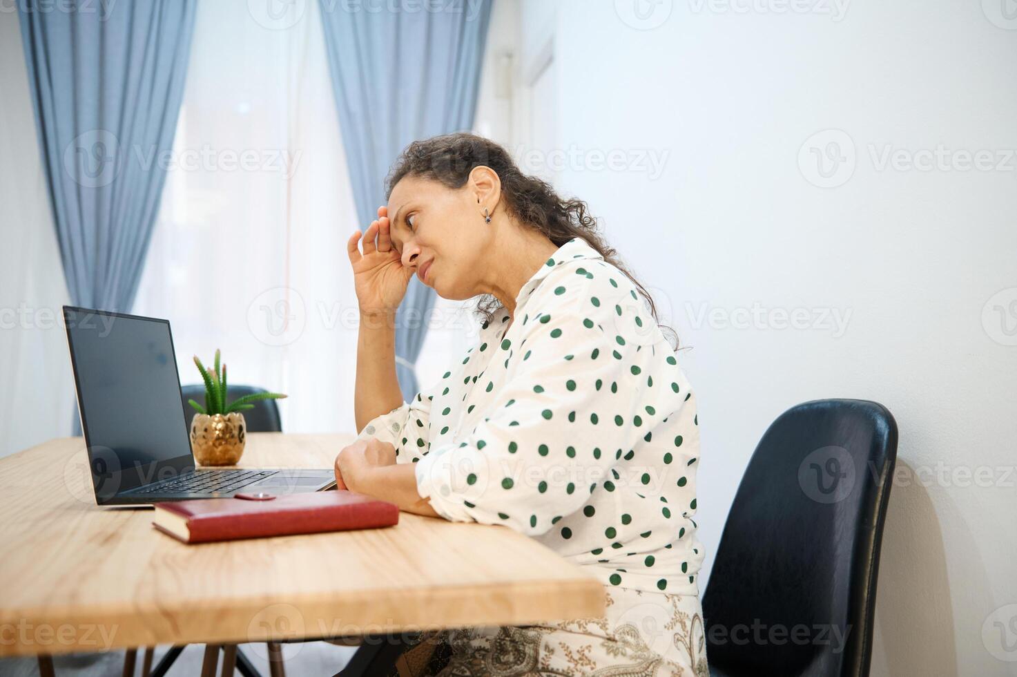 Woman in polka dot blouse looking stressed while working on a laptop at home office setup photo