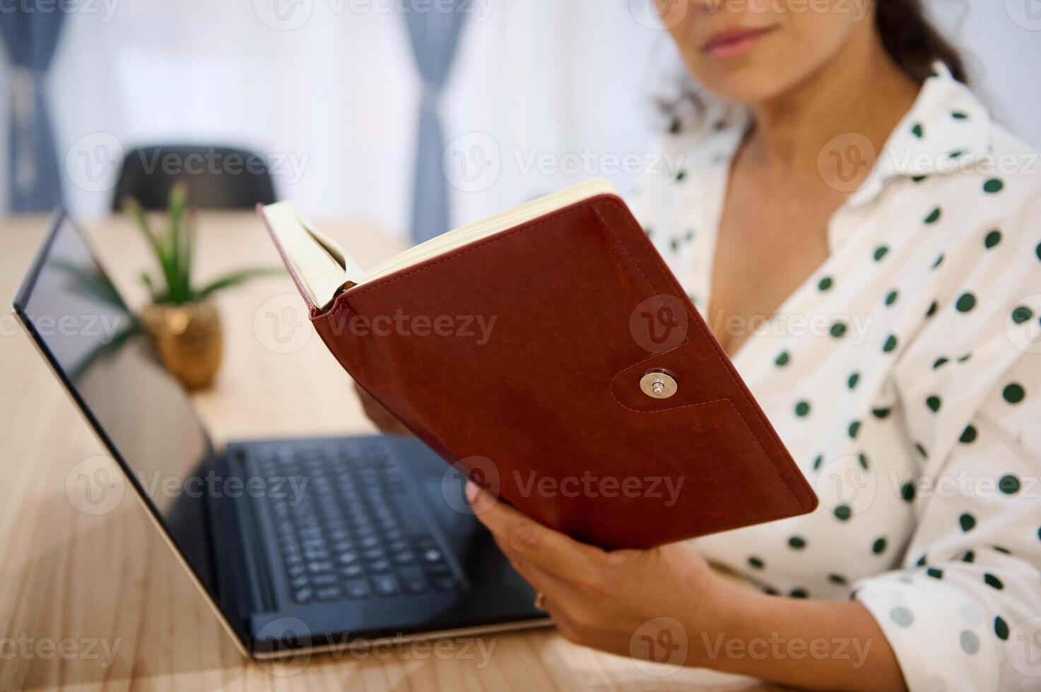 Woman in polka dot blouse reading a book at a table with a laptop open beside her, emphasizing education and relaxation. photo