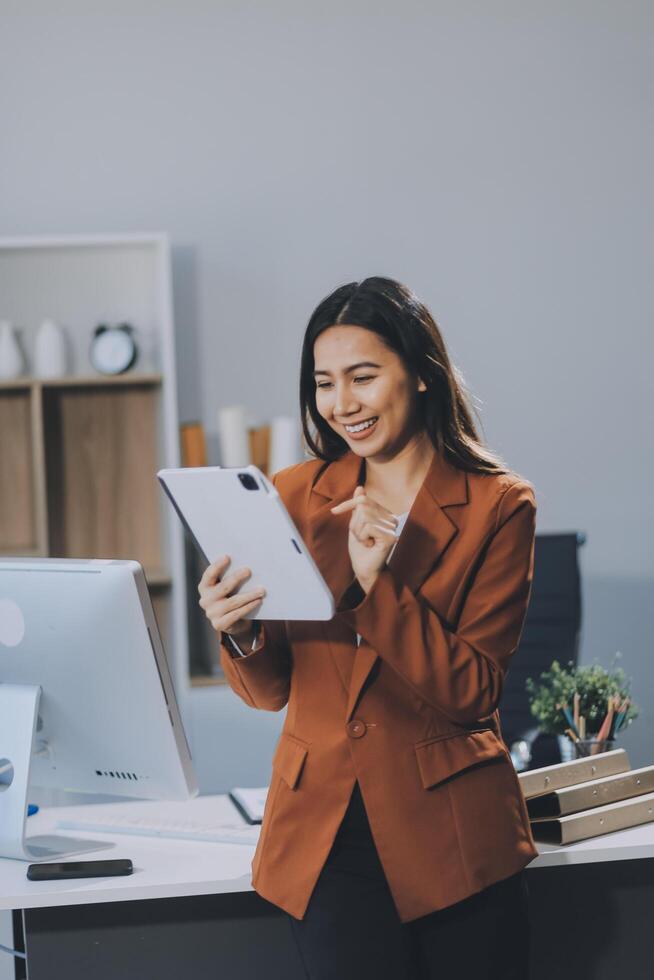 Attractive smiling Asian businesswoman standing holding tablet working and recording work details in office. photo