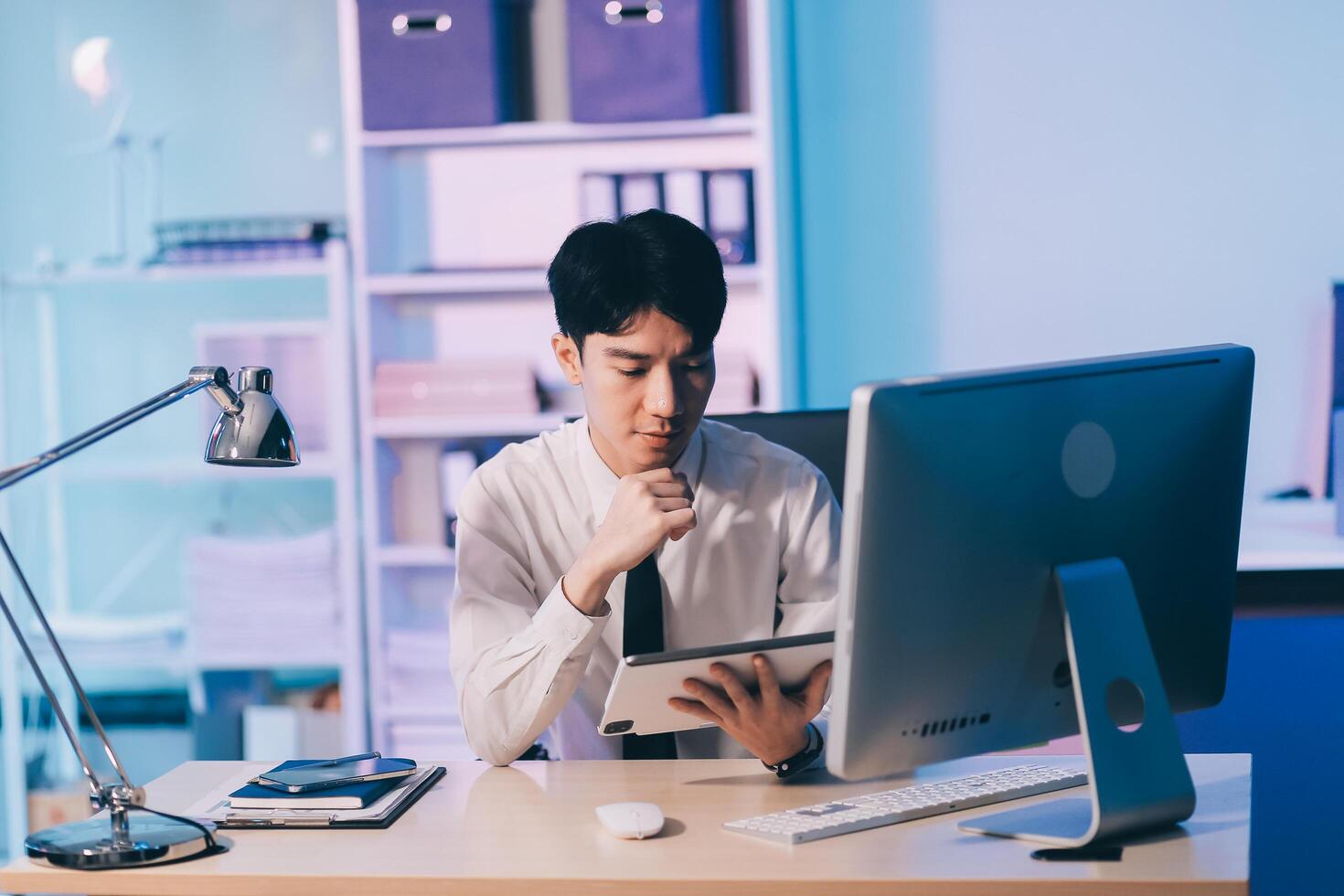 Young pensive asian man working late concentrated and serious in front of computer at night in dark office, Late night working or studying concept. photo