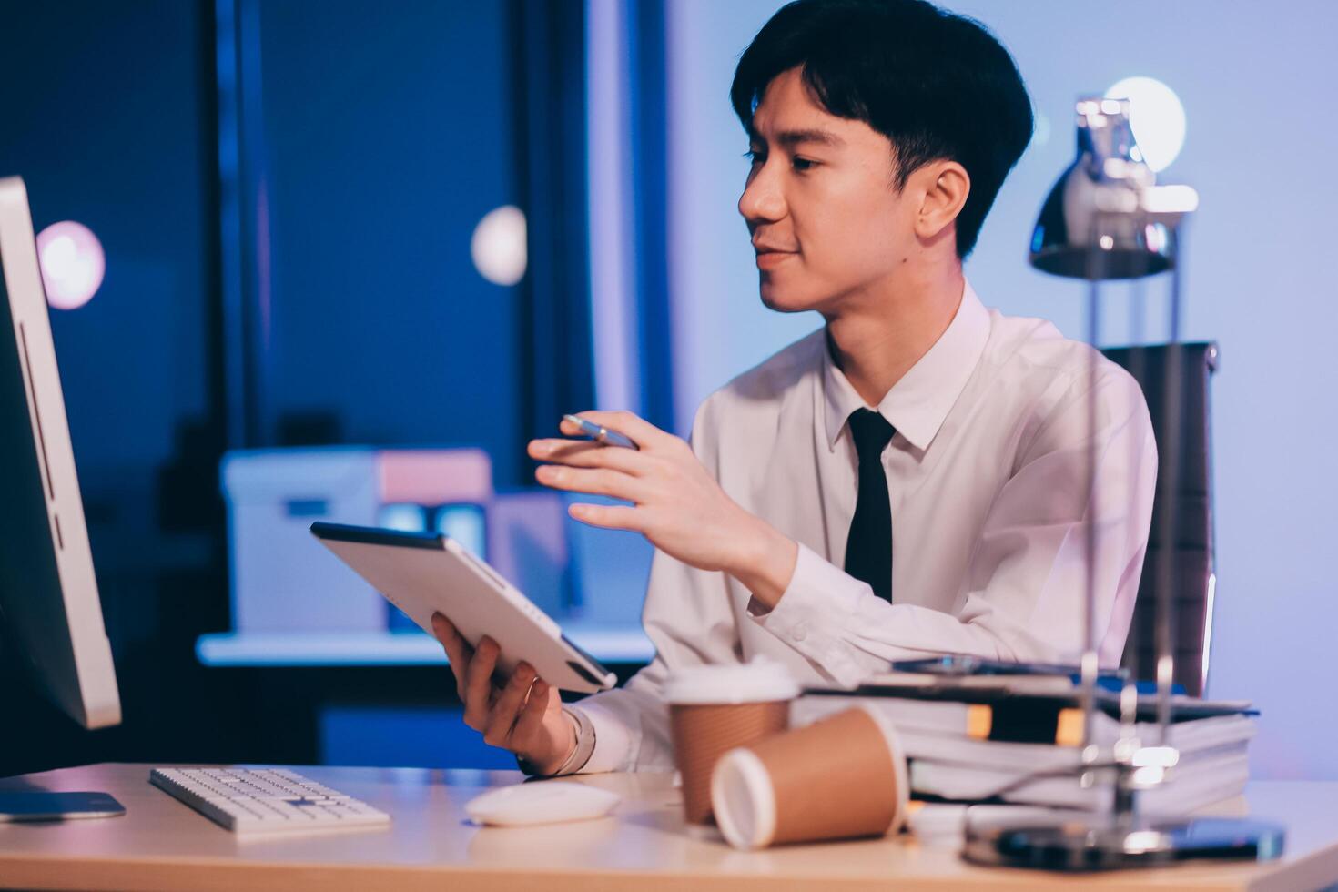 Young pensive asian man working late concentrated and serious in front of computer at night in dark office, Late night working or studying concept. photo