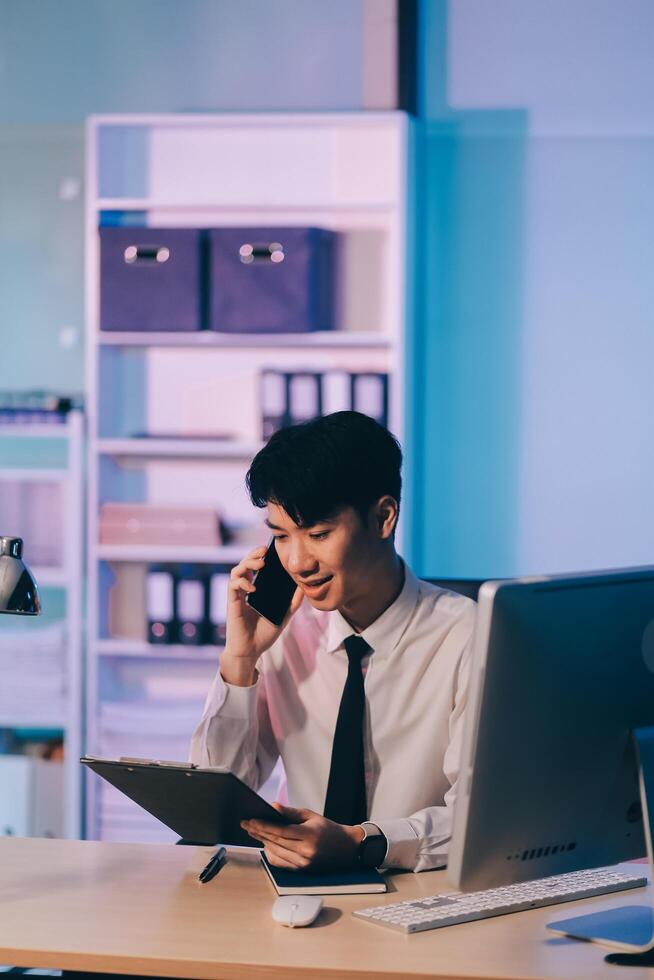 Young pensive asian man working late concentrated and serious in front of computer at night in dark office, Late night working or studying concept. photo
