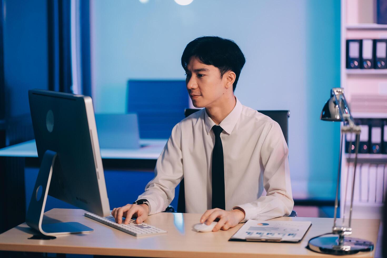 Young pensive asian man working late concentrated and serious in front of computer at night in dark office, Late night working or studying concept. photo