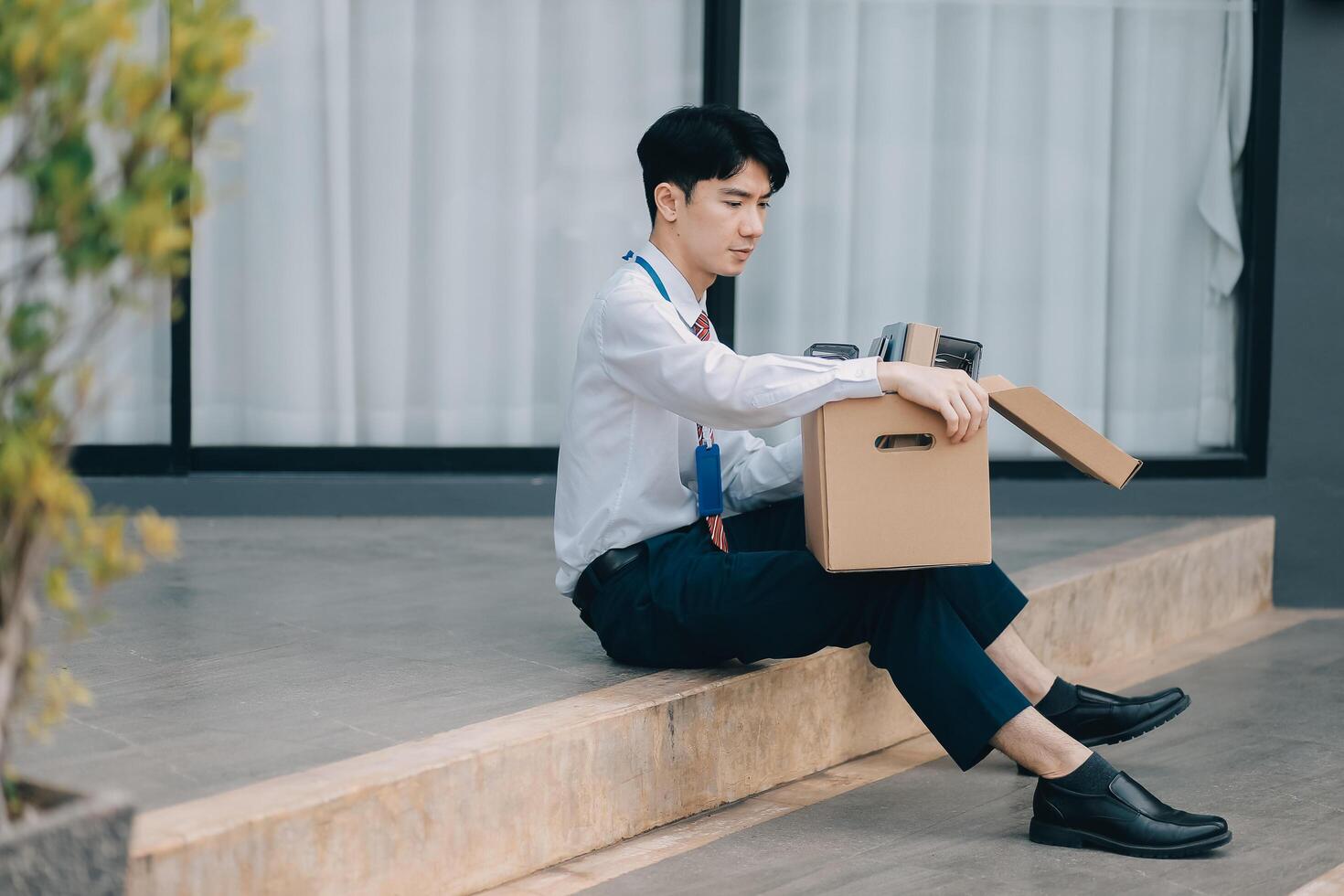 Sadness depressed Businessman with box cardboard packing personal items after losing jobs. Failure businessman sitting at stair front of building. Your fired Unemployed Jobless People Crisis photo