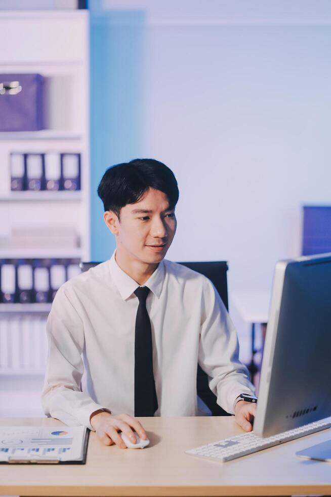 Young pensive asian man working late concentrated and serious in front of computer at night in dark office, Late night working or studying concept. photo