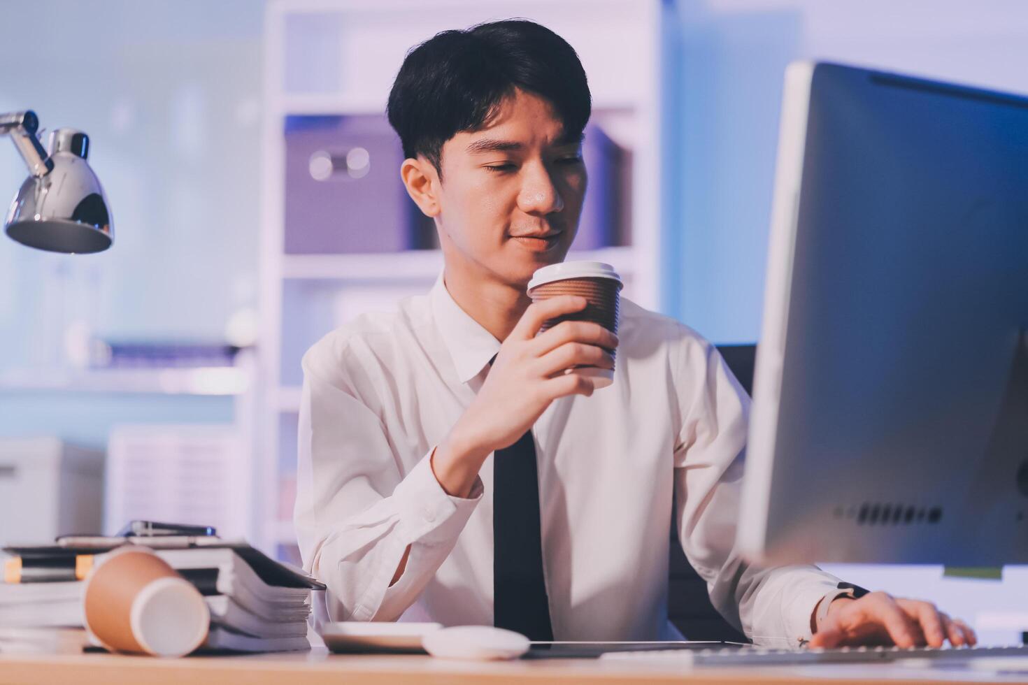 Young pensive asian man working late concentrated and serious in front of computer at night in dark office, Late night working or studying concept. photo
