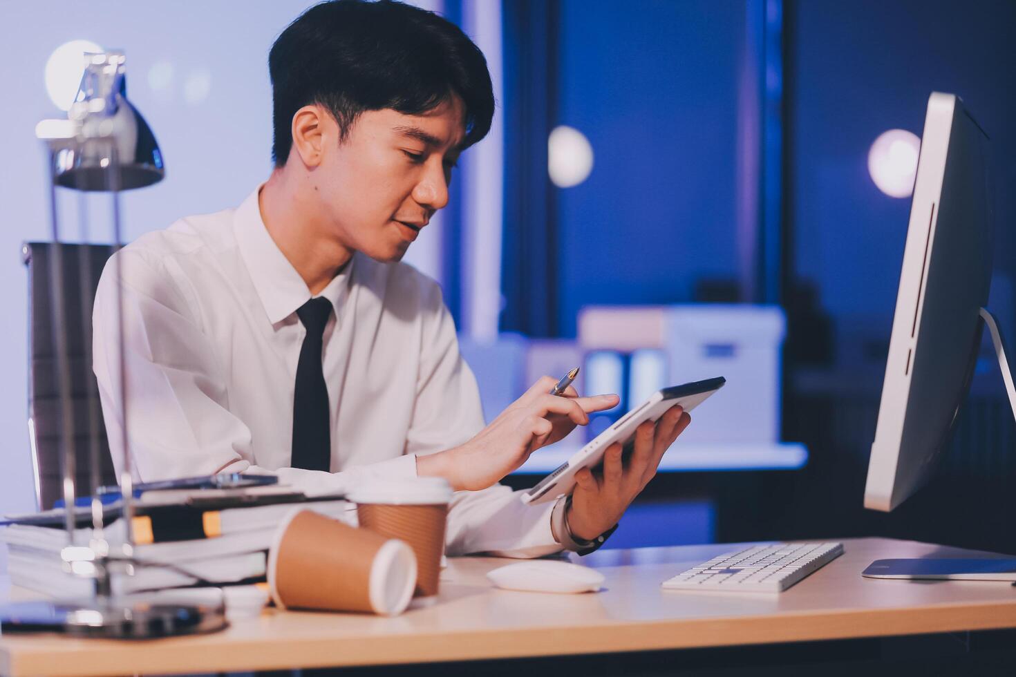 Young pensive asian man working late concentrated and serious in front of computer at night in dark office, Late night working or studying concept. photo