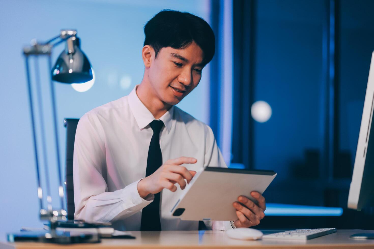 Young pensive asian man working late concentrated and serious in front of computer at night in dark office, Late night working or studying concept. photo