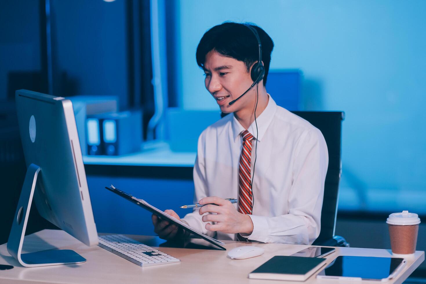 Happy smiling Asian man work at call center service desk consultant with teammates at night, customer service executive with microphone headset use computer for supporting in late night office. photo