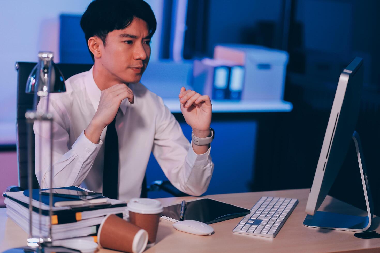 Young pensive asian man working late concentrated and serious in front of computer at night in dark office, Late night working or studying concept. photo