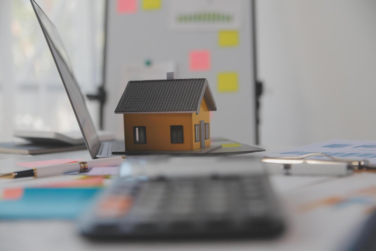 Wooden table with laptop, house keys Clipboard with pen, house model and green tree calculator, real estate concept. close up pictures photo