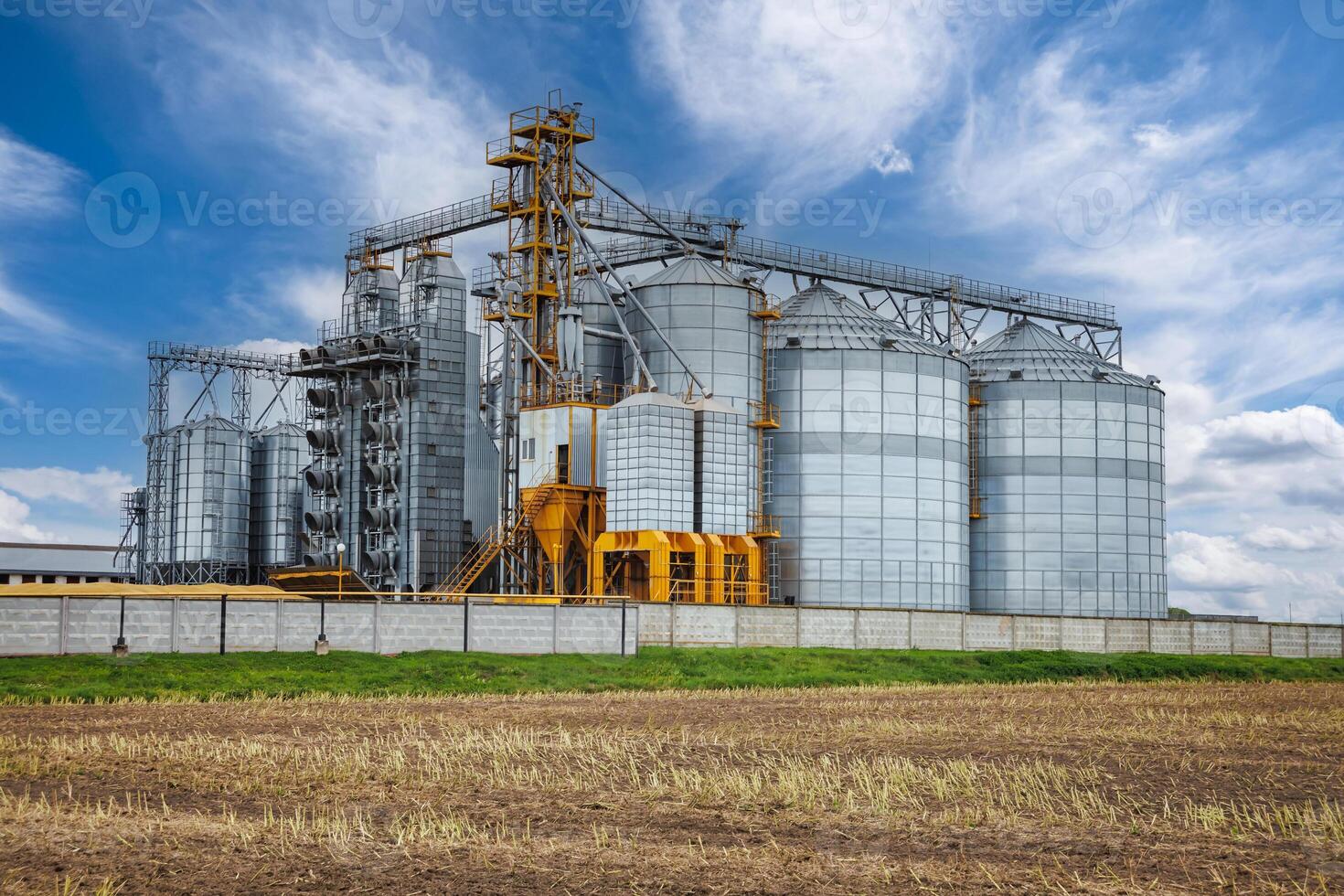 large grain elevator with multiple cylindrical metal silos, structural elements, and a complex system of pipes. It stands against a blue sky, with a field in the foreground after harvest photo