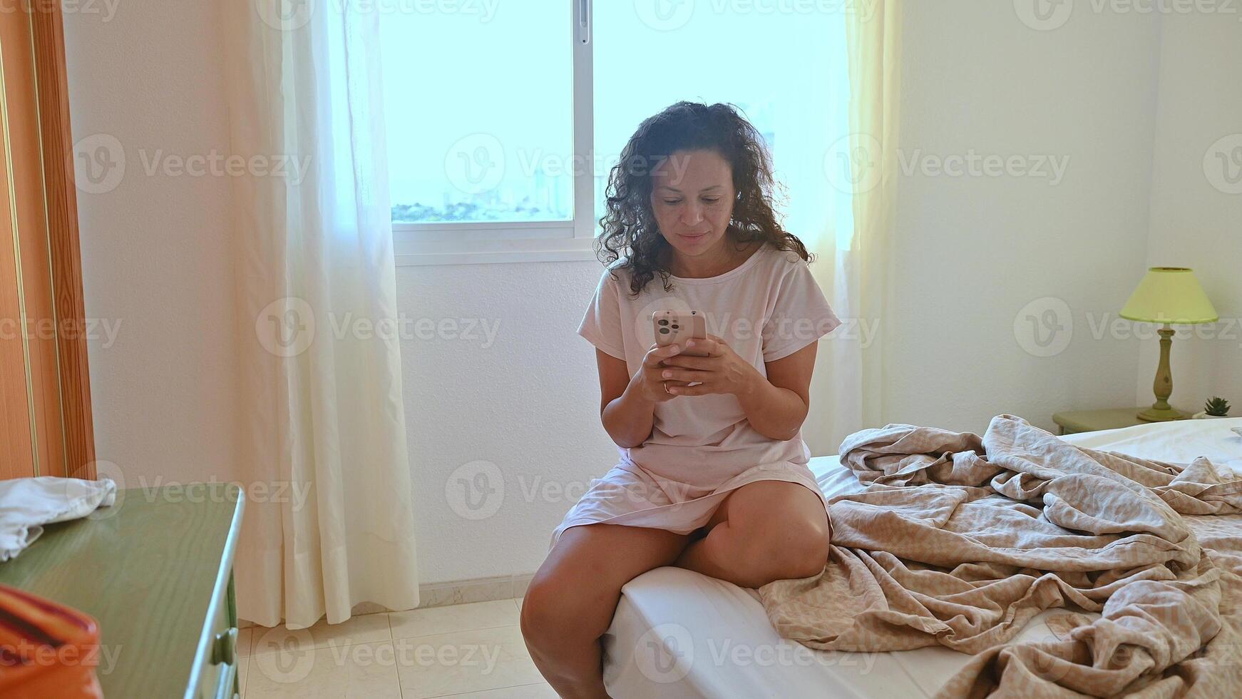 Woman in casual dress sitting on bed and using smartphone in cozy bedroom with natural light coming through the window photo