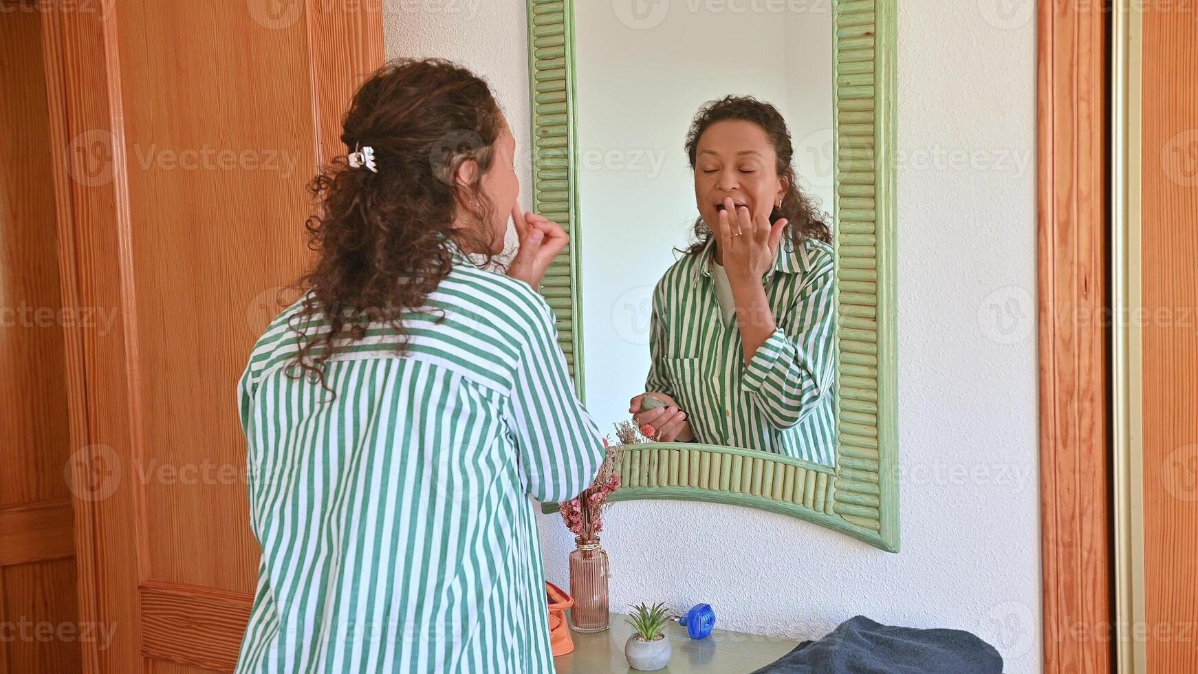 Woman in striped shirt applying moisturizer while looking in mirror at home for morning skincare routine photo
