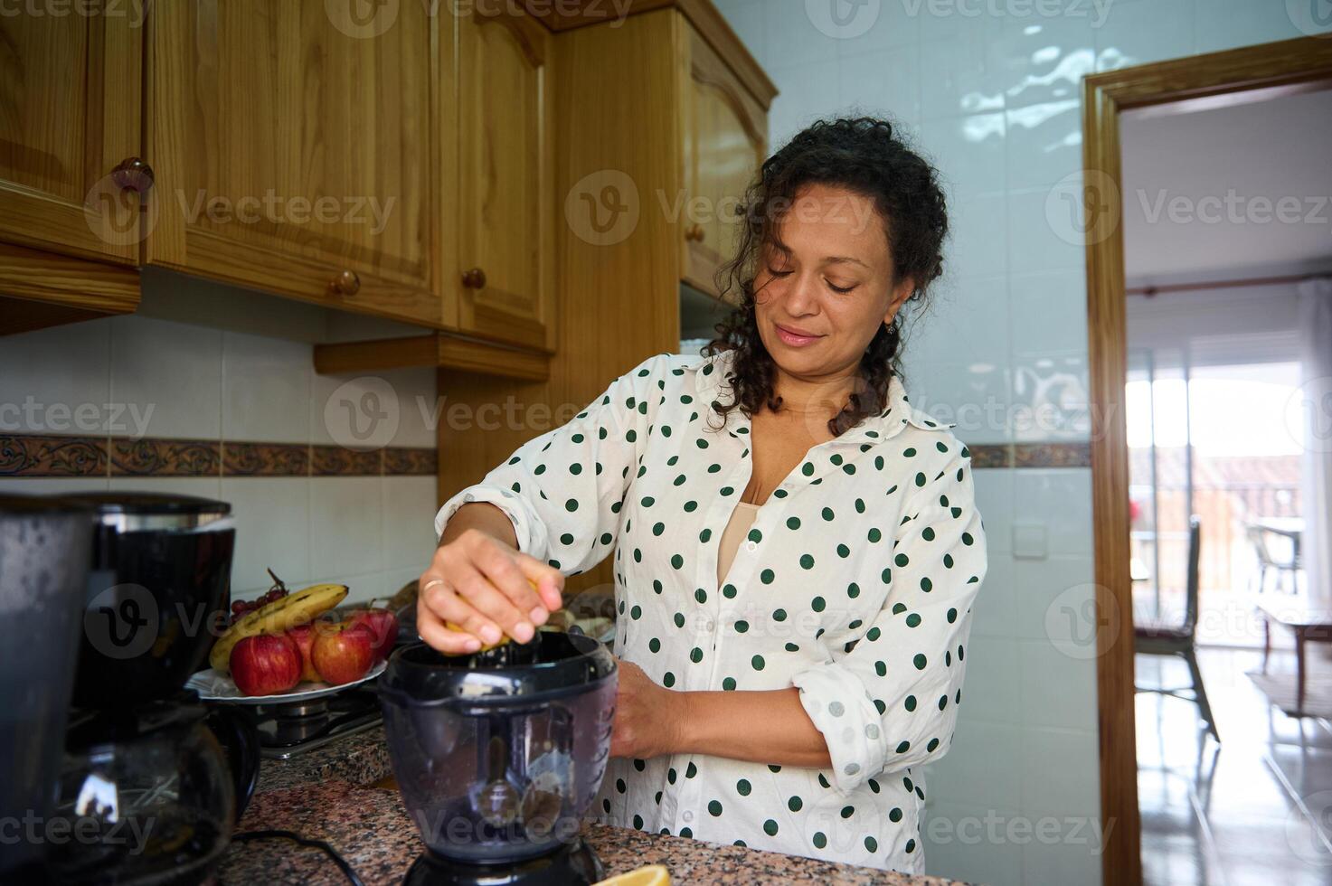 Woman in polka dot blouse using a blender in a cozy kitchen with fresh fruits on the counter, enjoying a relaxed morning routine photo