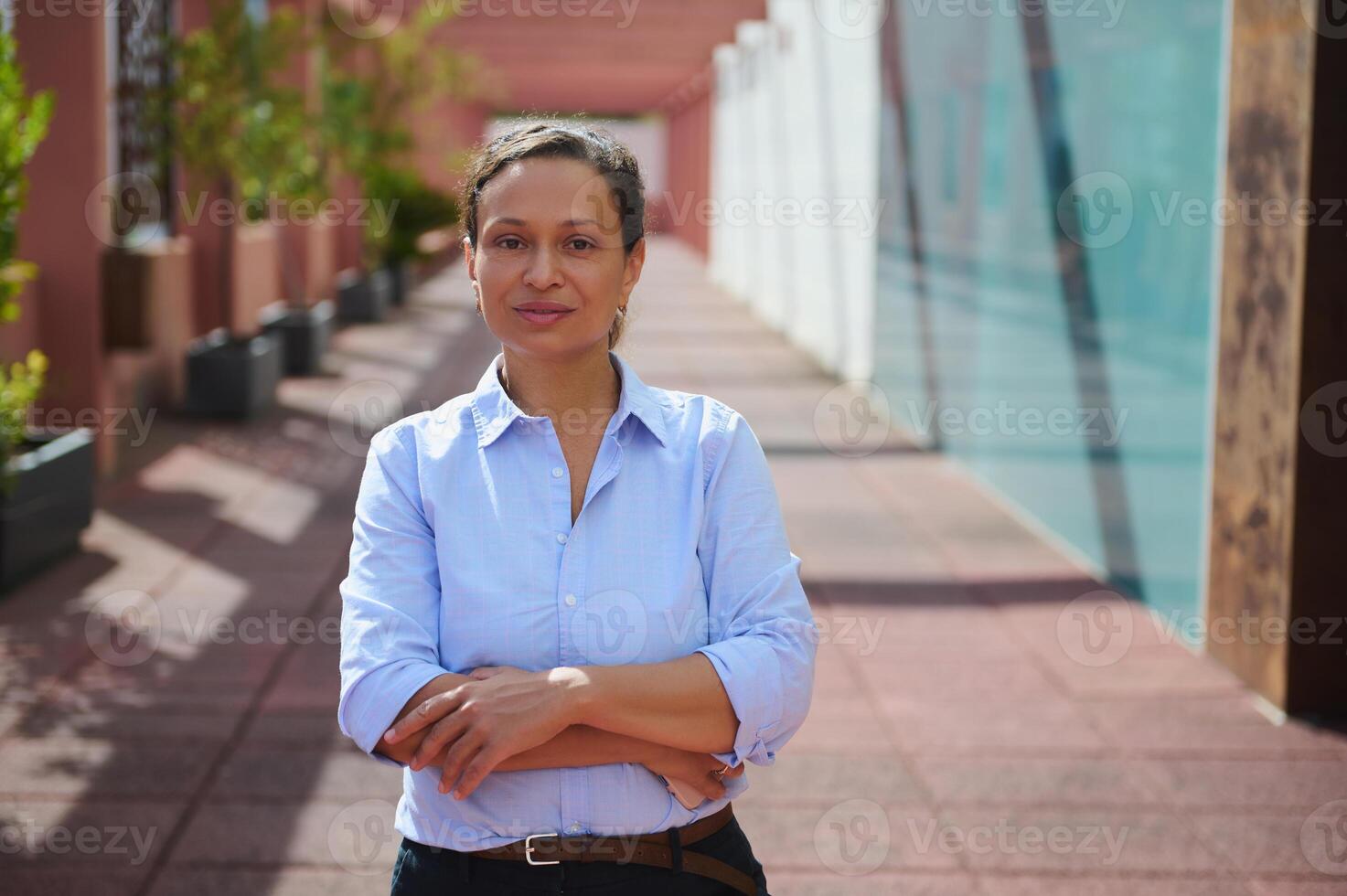 Confident professional woman standing outdoors with arms crossed in business attire, urban setting with modern architecture photo