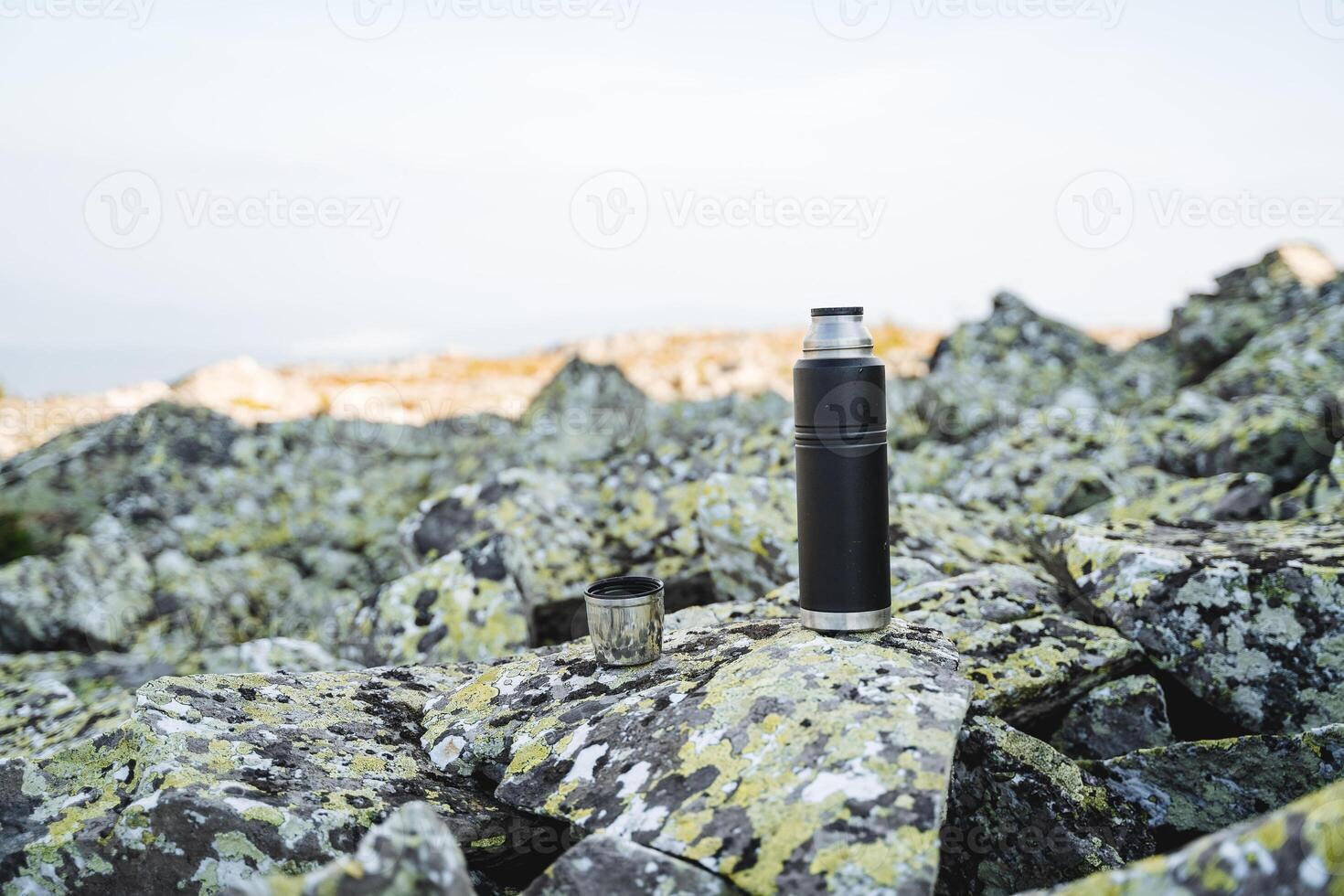 A thermos with tea stands on a stone, tourist dishes, a flask for hot drinks, a thermal mug, blocks of stone, brewed tea, a black thermos stands on a stone. photo