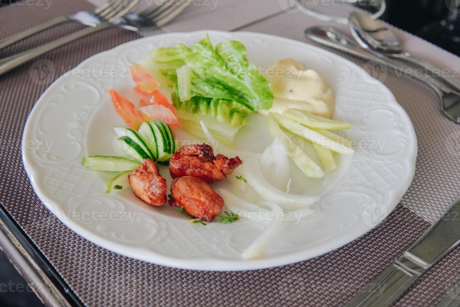 A plate of salad with a variety of vegetables and meat, set on a table with silverware. photo