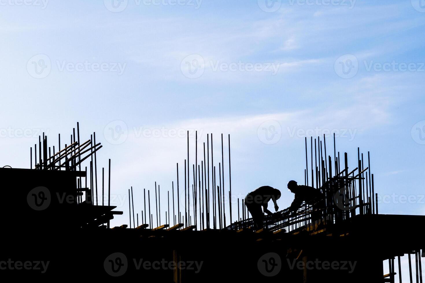 unrecognizable workers are working at construction site with many vertical rebars building a structure in front of blue sky in silhouettes photo