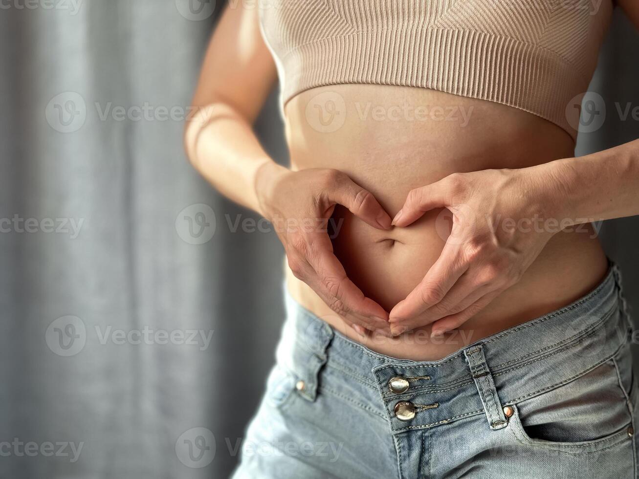 Close up of slim woman in short top and jeans forming heart shape with her hands on her bare stomach. Concept of body positivity, self love, and health awareness. Lifestyle photo. photo