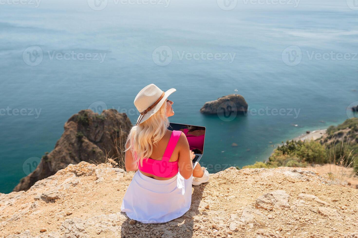 A woman is sitting on a rocky beach with a laptop in front of her. She is wearing a pink top and a straw hat. The beach is near the ocean, and the sky is clear. photo