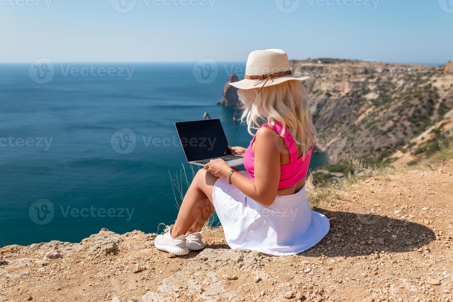 A woman is sitting on a rocky beach with a laptop open in front of her. She is wearing a pink top and a straw hat. Concept of relaxation and leisure. photo