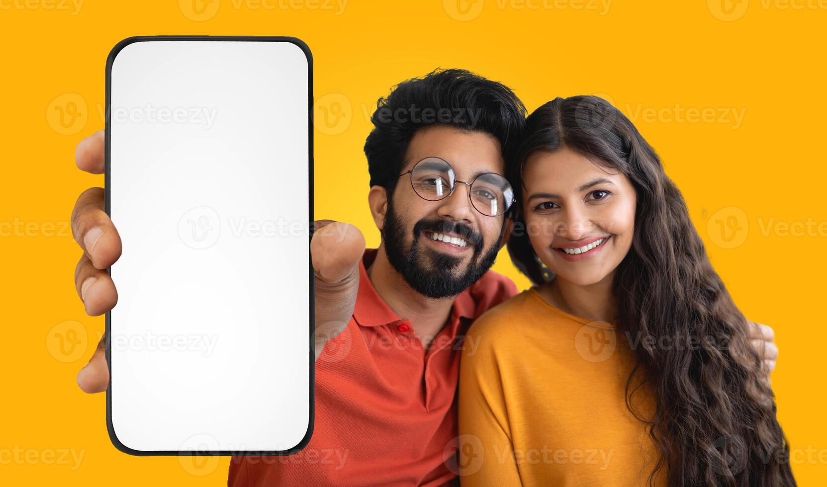 A young Indian couple, both with dark hair and smiles, are posing against a yellow background. They are holding a smartphone with a blank screen, which is positioned in front of the camera. photo